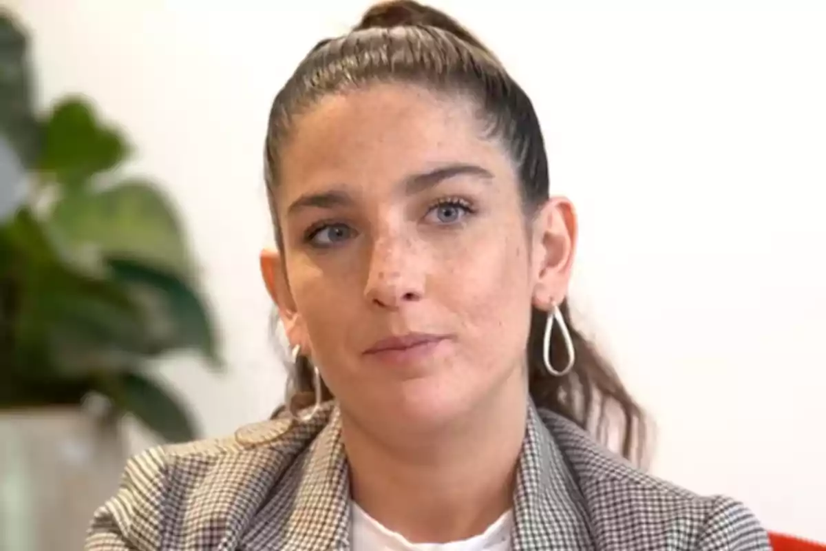 Una persona con cabello recogido y aretes grandes en un entorno interior con una planta al fondo.