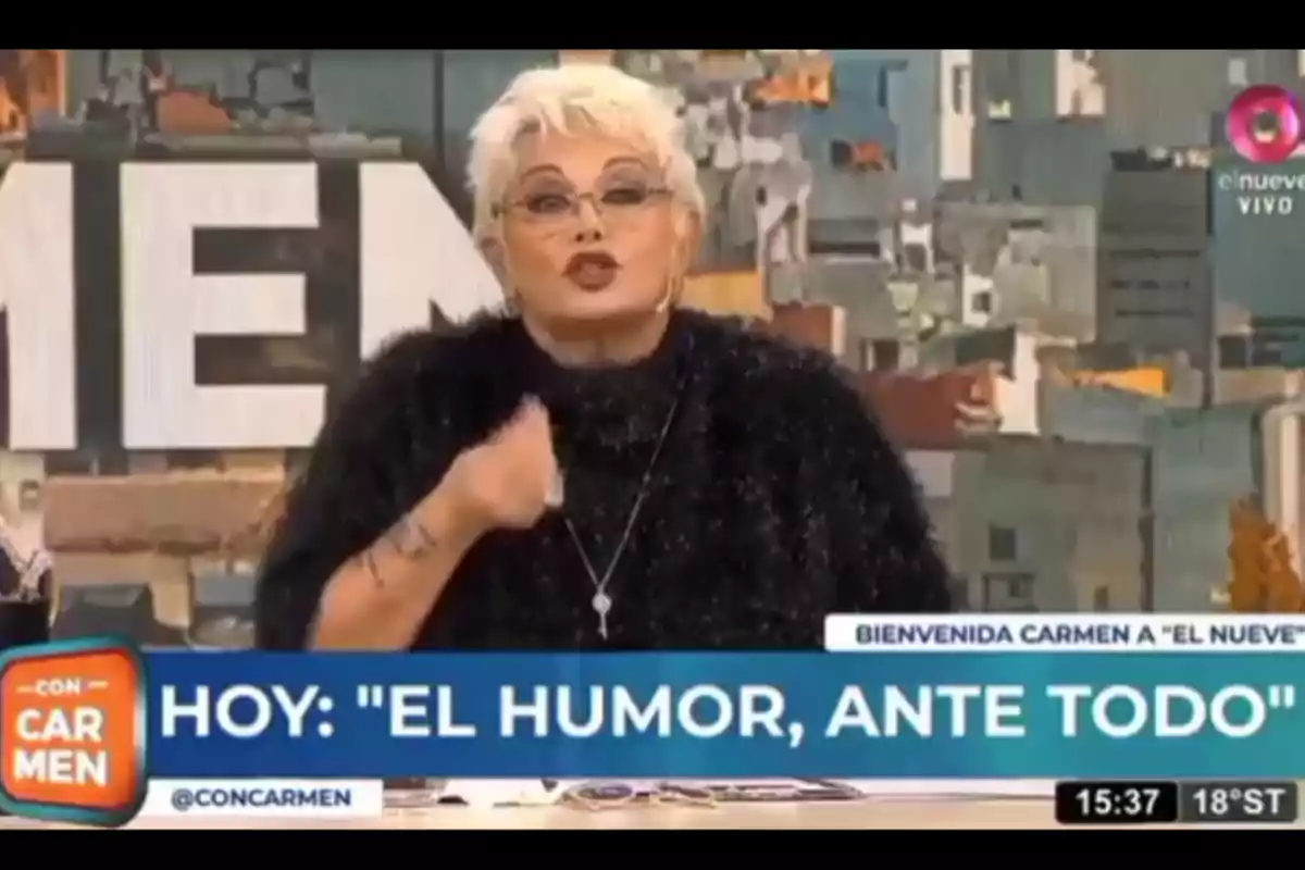 Mujer de cabello corto y rubio con gafas hablando en un programa de televisión, vistiendo un suéter negro y gesticulando con la mano, con un fondo de ciudad y gráficos en pantalla que anuncian el tema del día sobre el humor Mujer de cabello corto y rubio con gafas hablando en un programa de televisión, vistiendo un suéter negro y gesticulando con la mano, con un fondo de ciudad y gráficos en pantalla que anuncian el tema del día sobre el humor