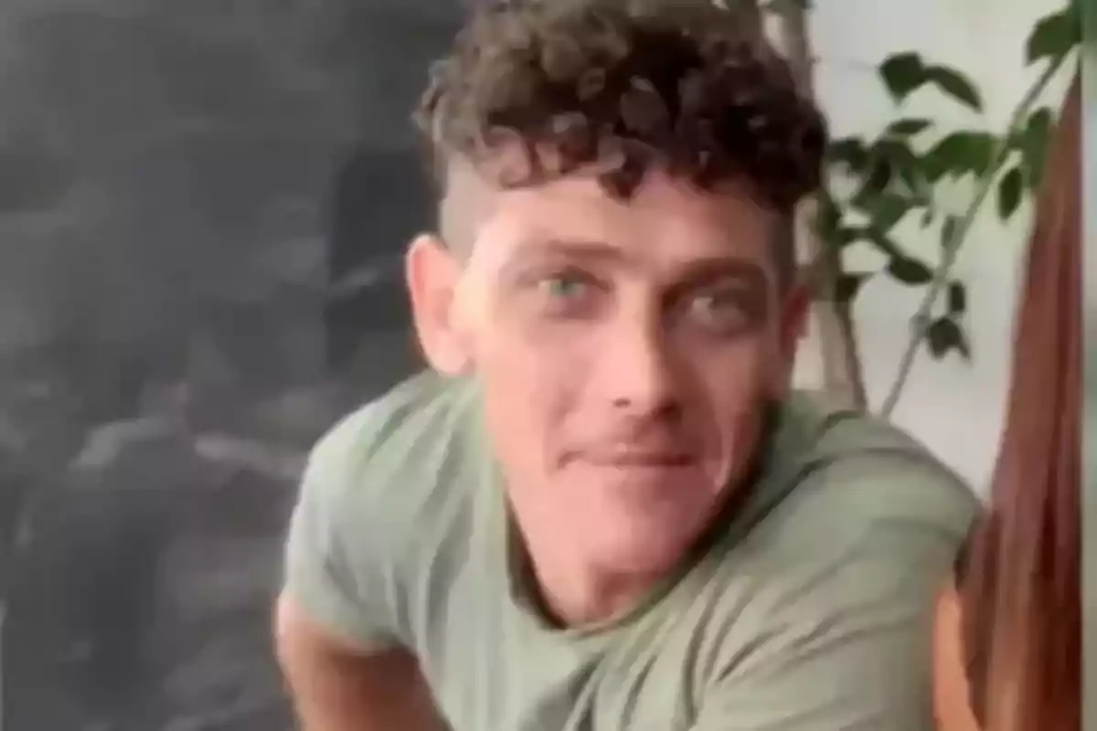 Curly-haired man with light eyes wearing a light green jersey sitting in front of a plant