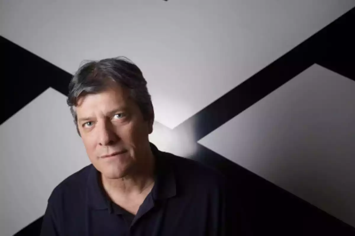 Hombre de cabello canoso y camisa oscura posando frente a una pared blanca con líneas negras en forma de equis