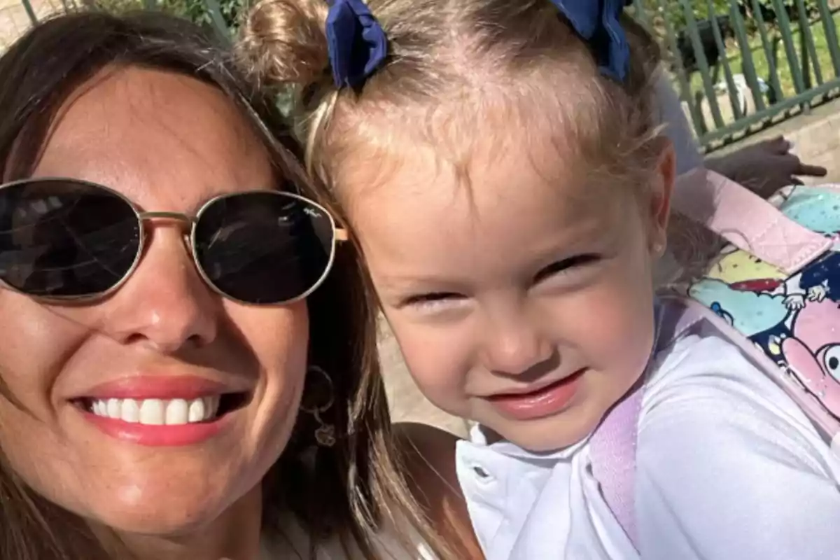 Una mujer con gafas de sol sonríe junto a una niña pequeña que lleva un lazo azul en el cabello.