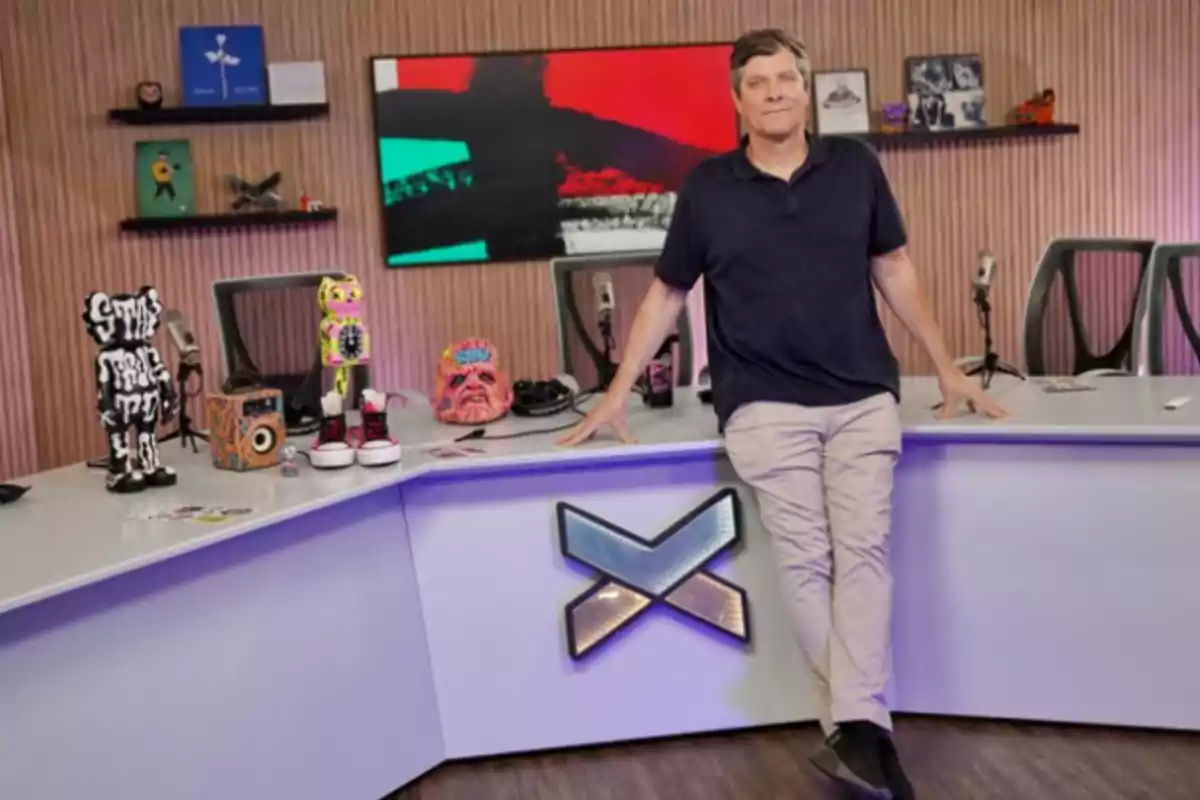 Hombre de pie junto a un escritorio moderno decorado con figuras y objetos coloridos en un estudio con fondo de madera y una pantalla con arte abstracto. Hombre de pie junto a un escritorio moderno decorado con figuras y objetos coloridos en un estudio con fondo de madera y una pantalla con arte abstracto.