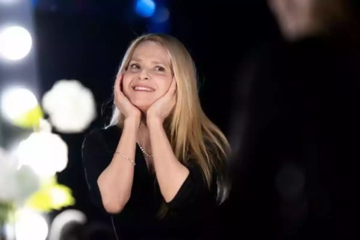 Mujer rubia sonriente con las manos en las mejillas mirando hacia arriba en un ambiente iluminado y elegante