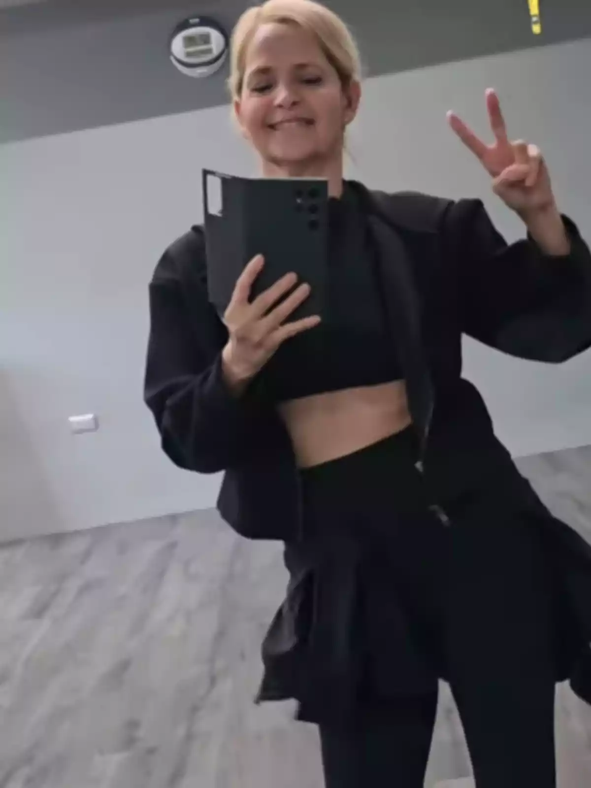 Mujer rubia sonriente tomándose una selfie frente a un espejo mientras hace el signo de la paz con la mano en un salón de piso de madera Mujer rubia sonriente tomándose una selfie frente a un espejo mientras hace el signo de la paz con la mano en un salón de piso de madera