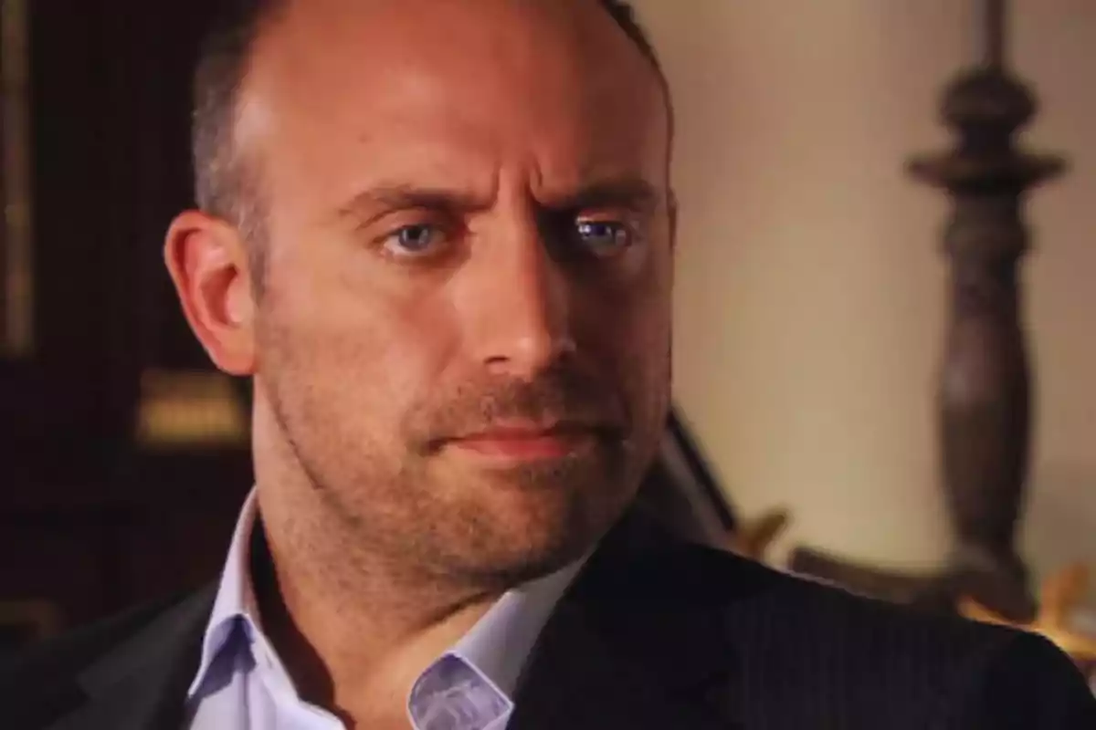 Bald man with a short beard and serious expression wearing a dark jacket and light jersey in an indoor setting