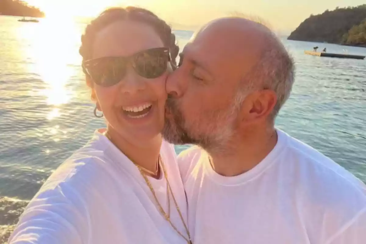 Smiling couple taking a selfie by the sea at sunset while the man kisses the woman on the cheek Smiling couple taking a selfie by the sea at sunset while the man kisses the woman on the cheek