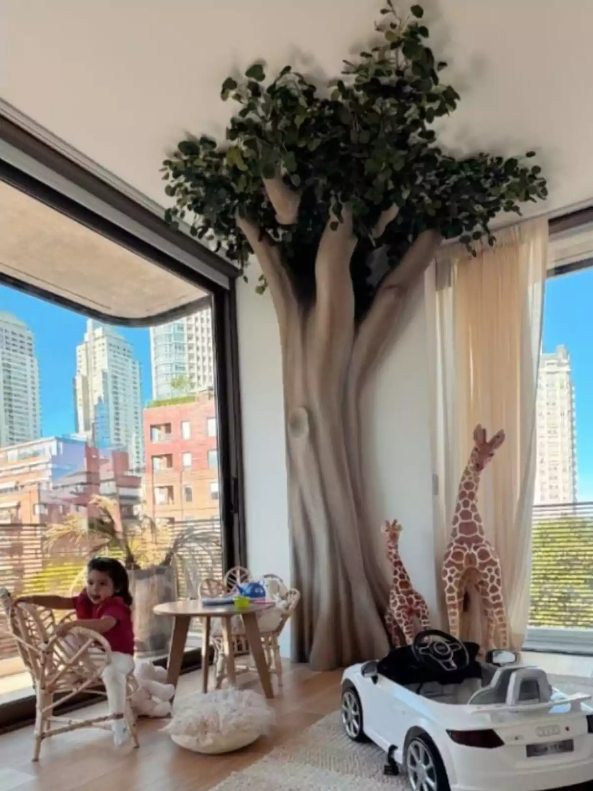 Una niña pequeña juega en una sala infantil decorada con un árbol artificial, dos jirafas de peluche y un auto de juguete blanco, con grandes ventanales que muestran edificios de la ciudad al fondo Una niña pequeña juega en una sala infantil decorada con un árbol artificial, dos jirafas de peluche y un auto de juguete blanco, con grandes ventanales que muestran edificios de la ciudad al fondo