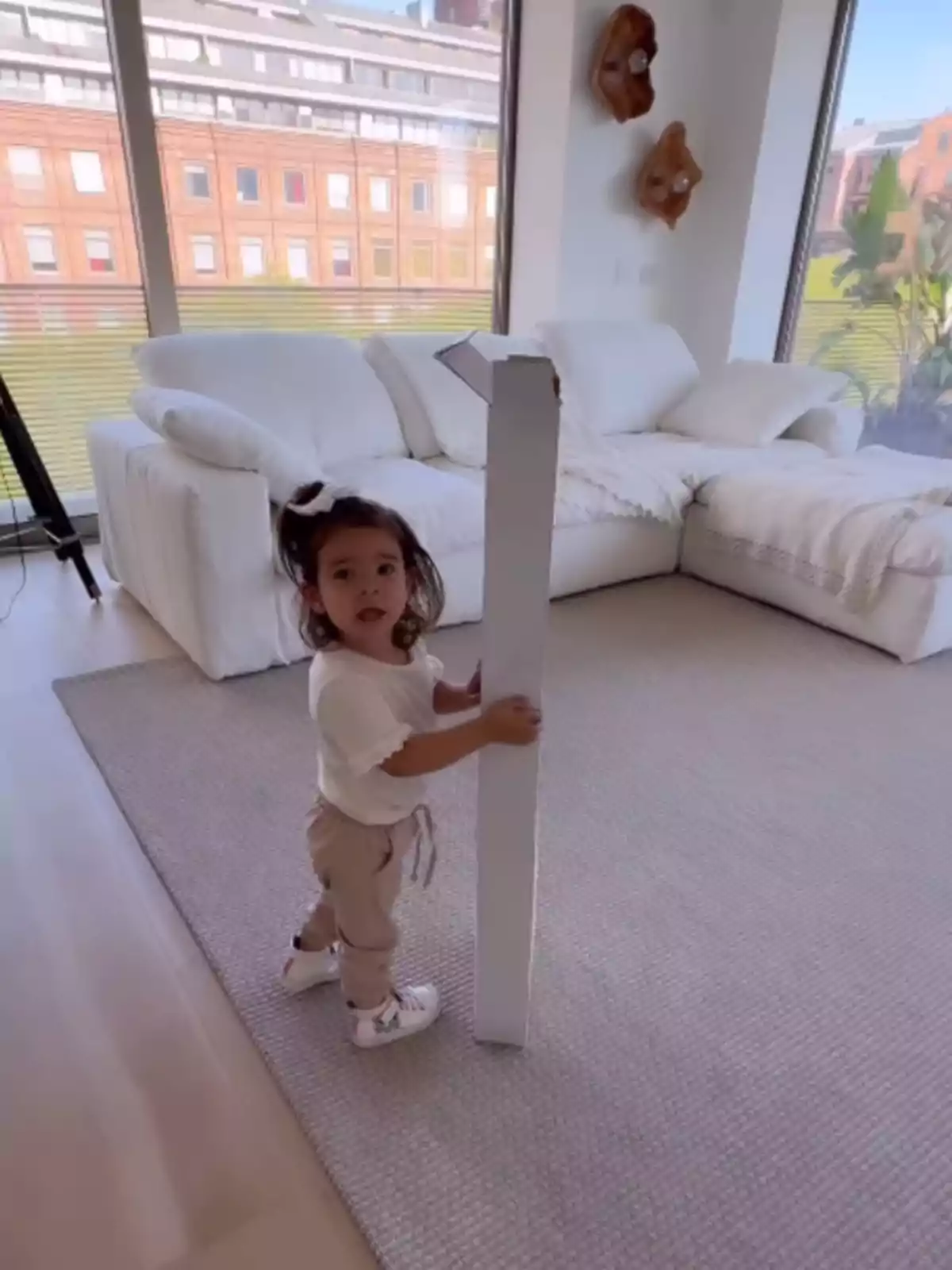 Niña pequeña de pie en una sala luminosa sosteniendo una caja larga de cartón frente a un sofá blanco Niña pequeña de pie en una sala luminosa sosteniendo una caja larga de cartón frente a un sofá blanco