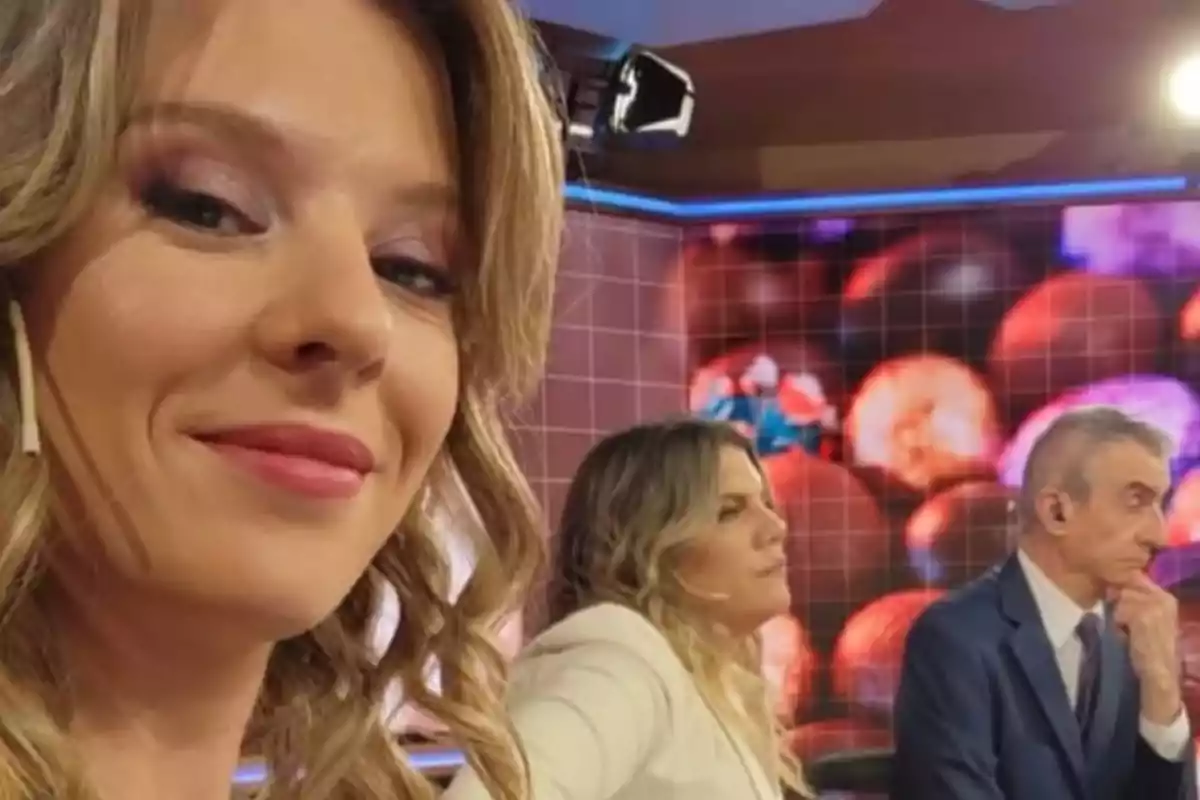 Tres personas en un estudio de televisión, una mujer en primer plano sonríe mientras al fondo una mujer y un hombre están sentados frente a una mesa
