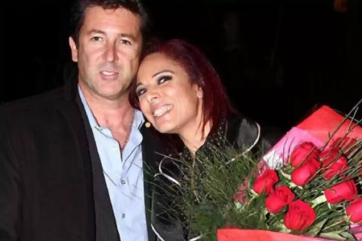 Una pareja sonríe mientras posan juntos, la mujer sostiene un ramo de rosas rojas Una pareja sonríe mientras posan juntos, la mujer sostiene un ramo de rosas rojas