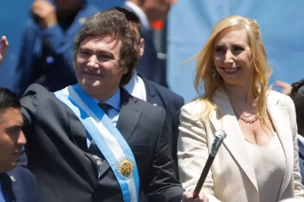 A man and a woman smile while standing at a formal event, the man wears a presidential sash and the woman holds a ceremonial staff.