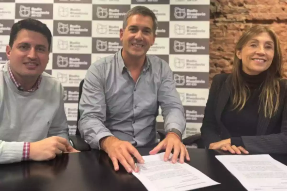 Three people sitting at a table smile as they pose in front of documents, with a background of repeated radio station logos. Three people sitting at a table smile as they pose in front of documents, with a background of repeated radio station logos.
