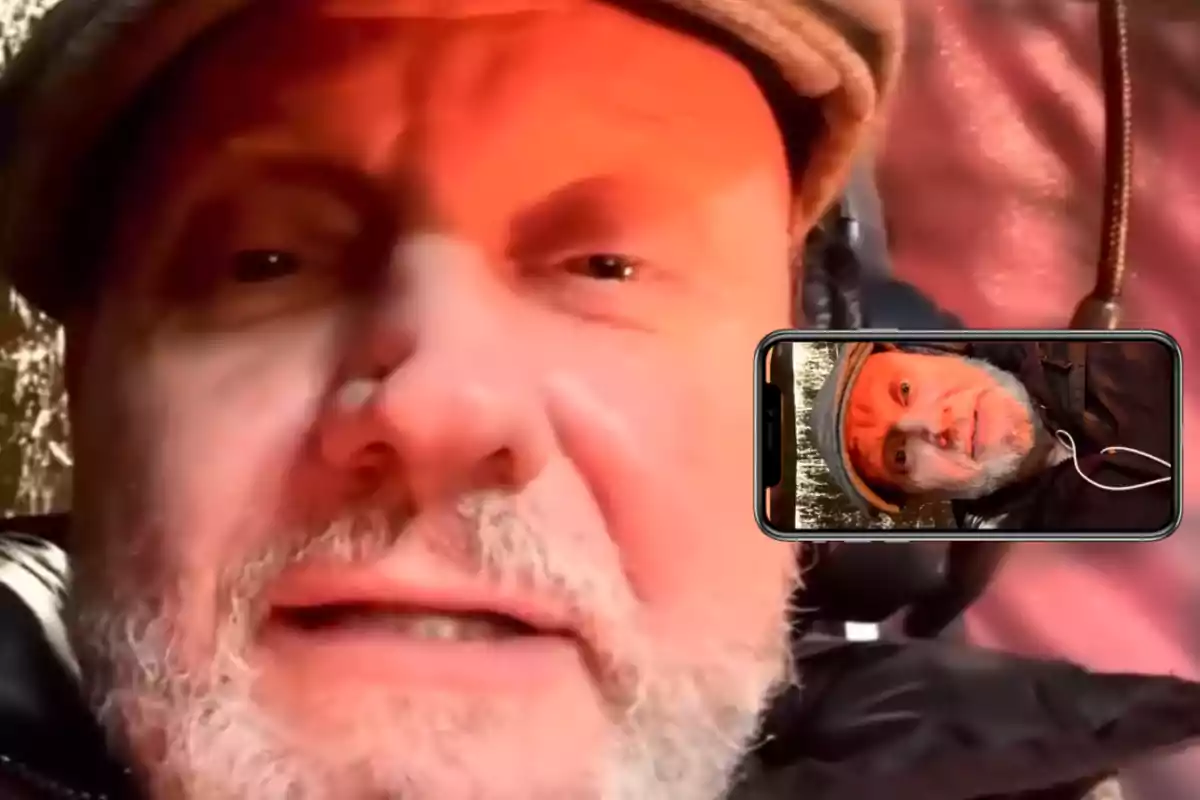 Gray-bearded man wearing a cap looking at the camera with a mobile phone showing his own face on a video call