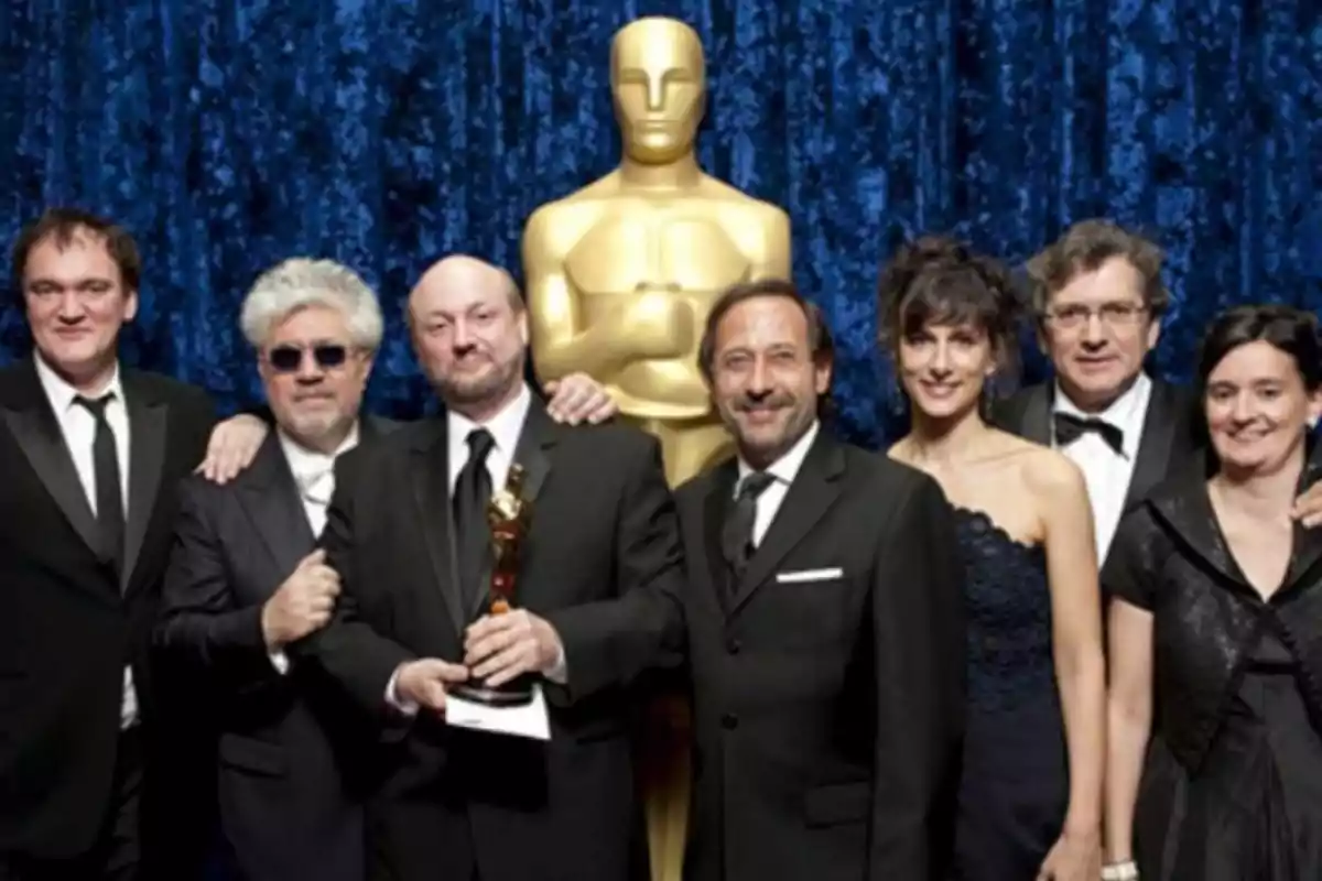 Group of elegantly dressed people posing in front of a large golden Oscar statue, one of them holding an award statuette. Group of elegantly dressed people posing in front of a large golden Oscar statue, one of them holding an award statuette.