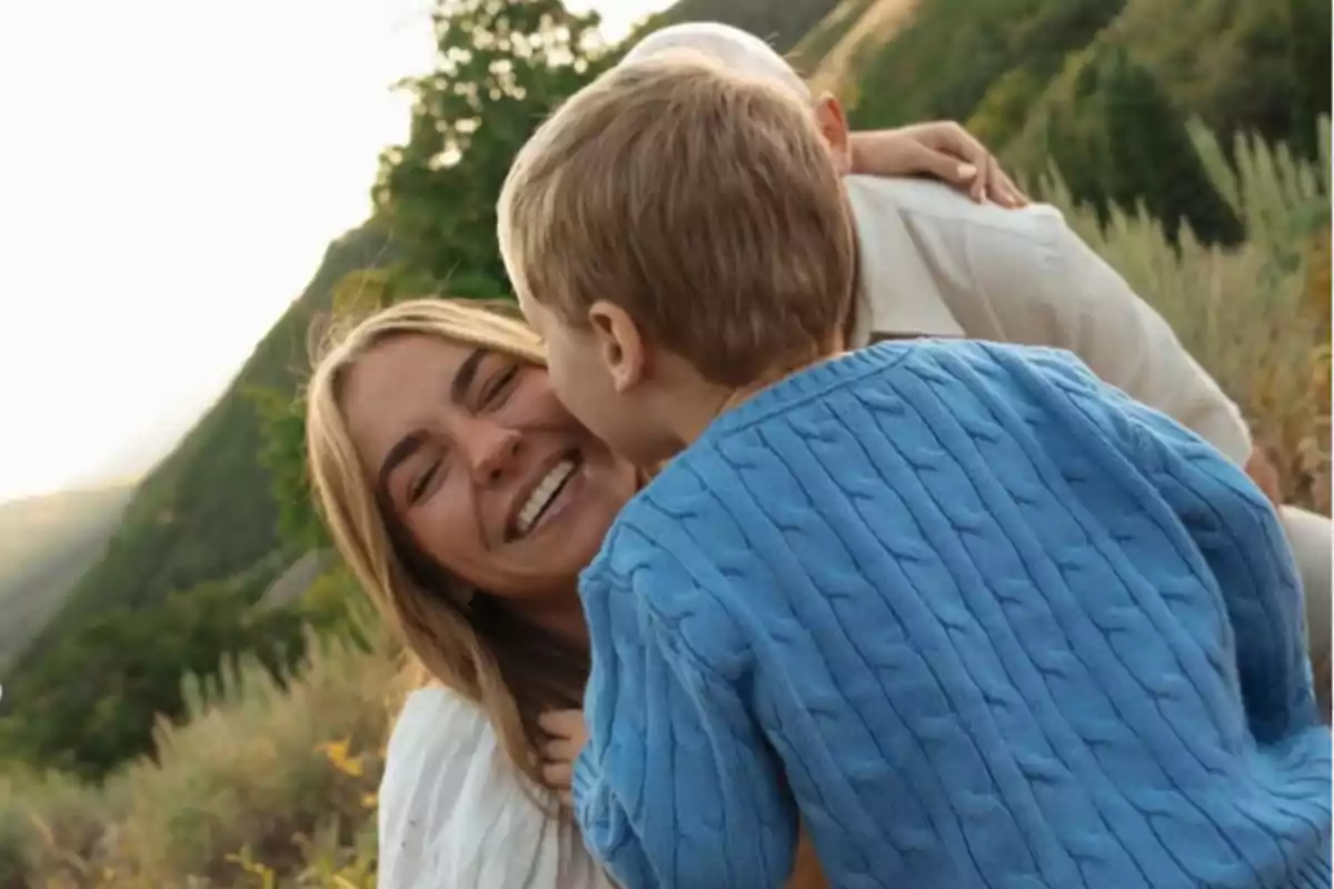 A woman smiles as a small child in a blue sweater gives her a kiss on the cheek in a natural setting.