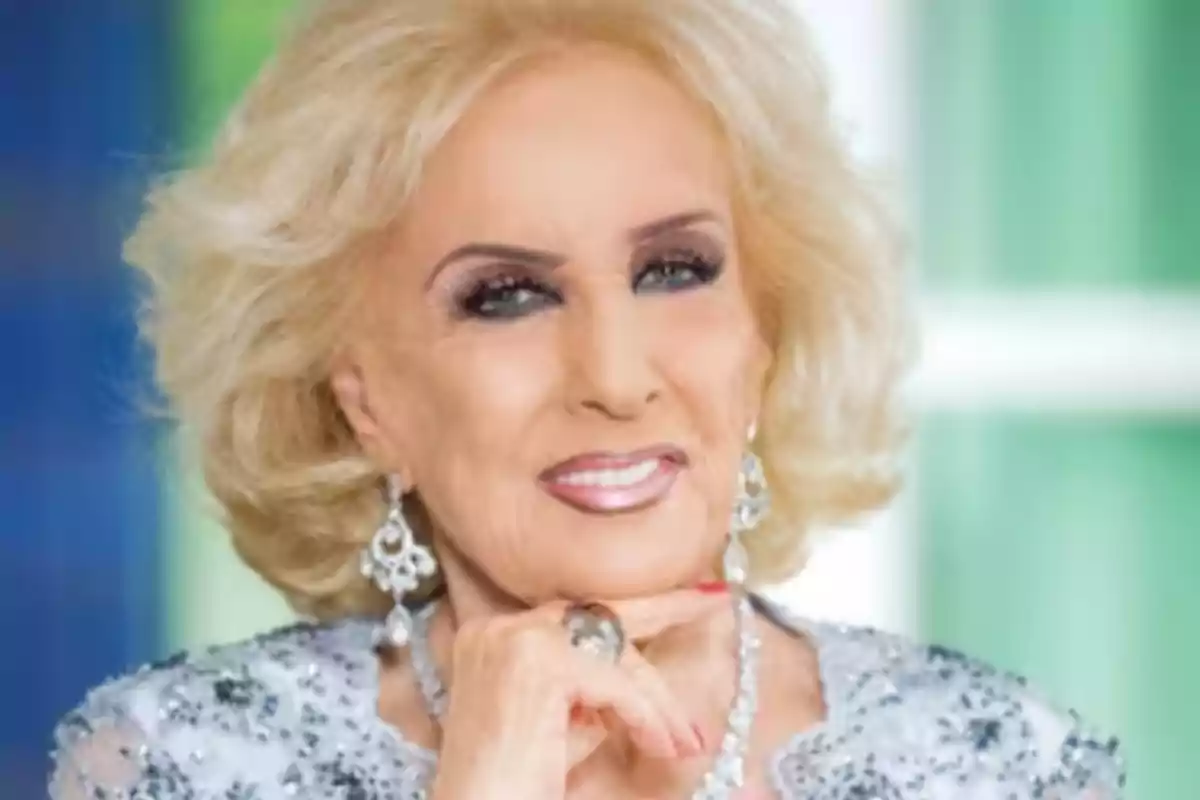 Older woman with short blonde hair, elegant makeup, wearing jewelry and a sparkling dress, smiling and resting her chin on one hand
