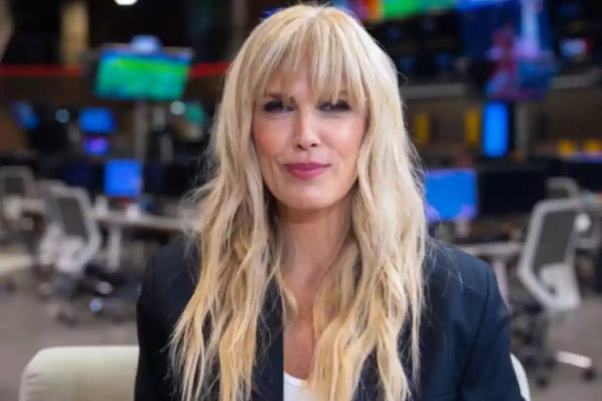 A person with long blonde hair is in an office setting with screens and desks in the background.