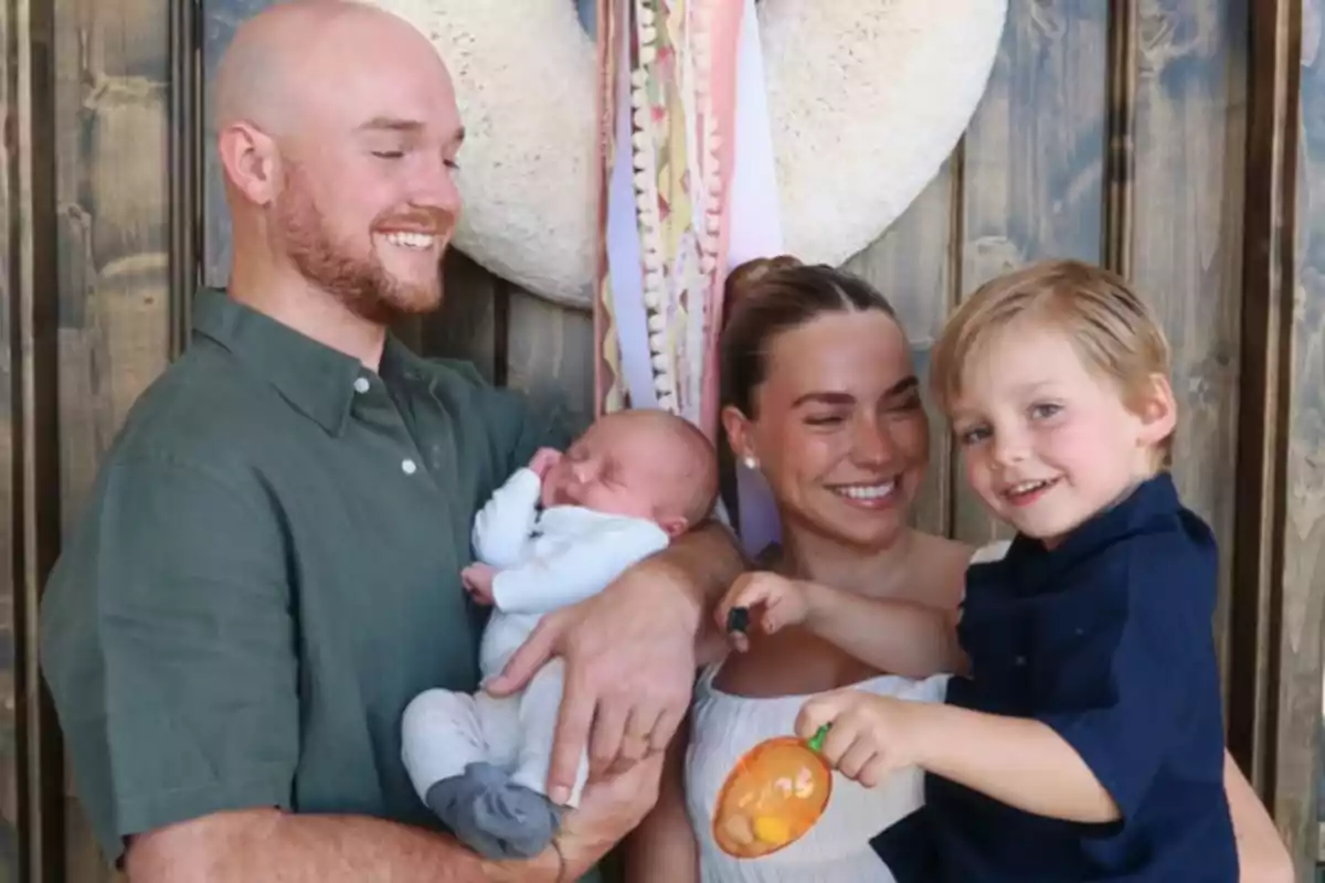 Smiling family posing together in front of a wooden wall, the father is holding a baby in his arms, the mother is smiling, and a young child is holding a toy. Smiling family posing together in front of a wooden wall, the father is holding a baby in his arms, the mother is smiling, and a young child is holding a toy.