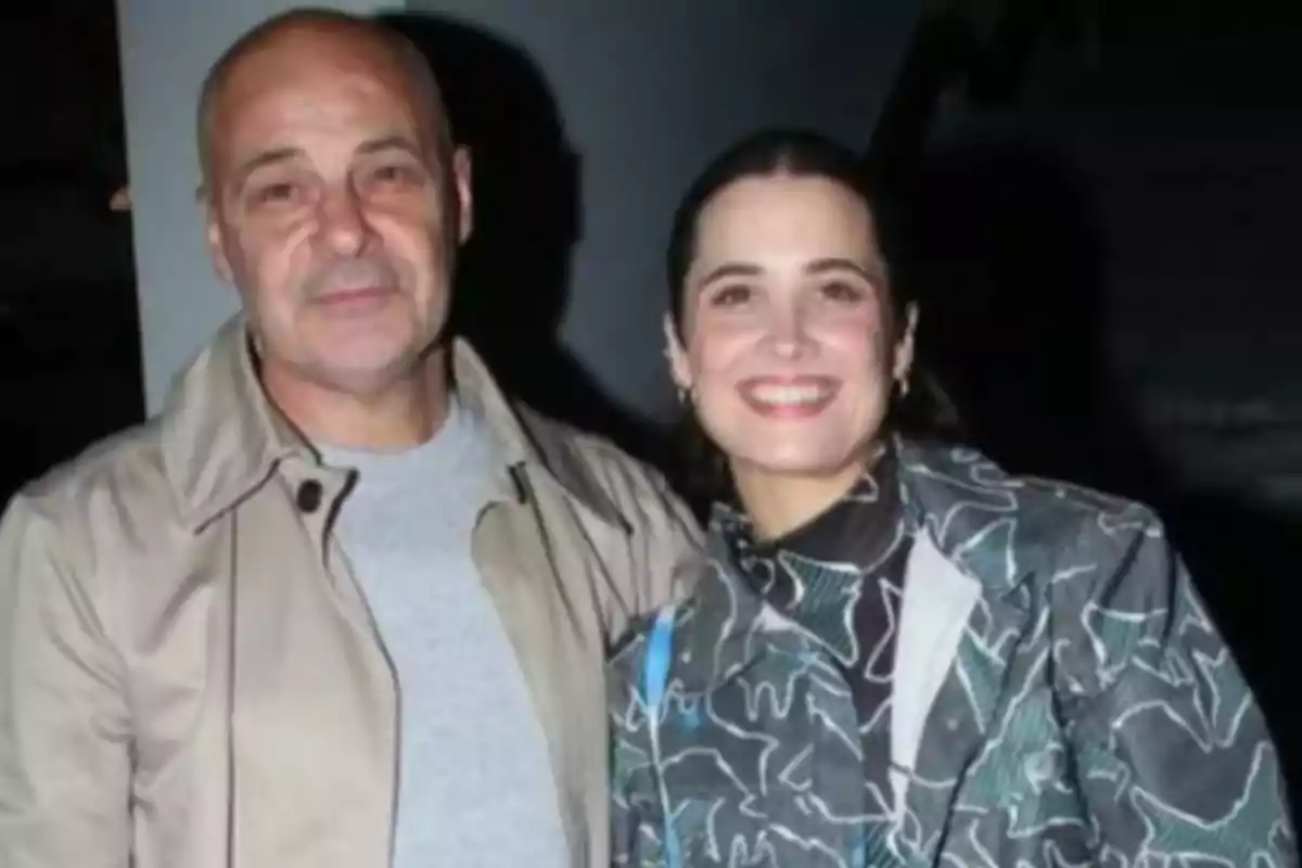 Una pareja sonriente posando para una foto, el hombre lleva una chaqueta beige y la mujer una chaqueta con diseño abstracto.