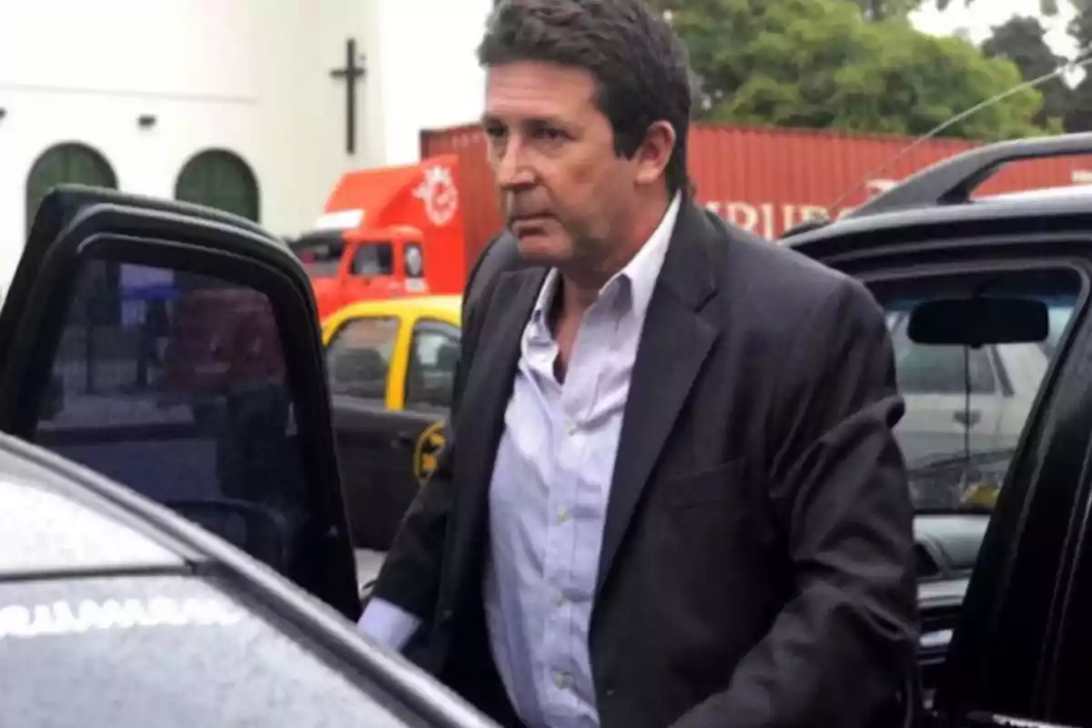 Hombre de cabello corto y canoso con saco oscuro y camisa clara bajando de un auto en la calle con vehículos y un edificio al fondo Hombre de cabello corto y canoso con saco oscuro y camisa clara bajando de un auto en la calle con vehículos y un edificio al fondo
