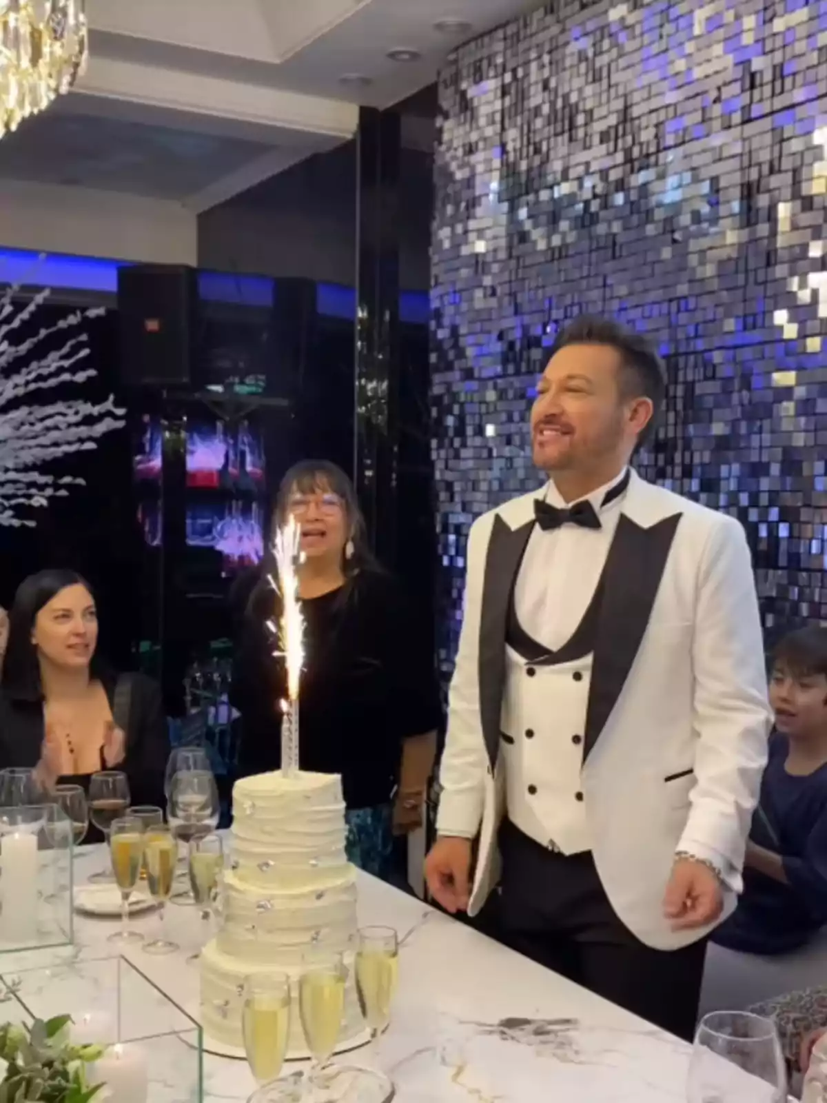 Hombre vestido de esmoquin blanco celebrando su cumpleaños frente a un pastel de varios pisos con una vela de chispas rodeado de personas en una mesa elegante Hombre vestido de esmoquin blanco celebrando su cumpleaños frente a un pastel de varios pisos con una vela de chispas rodeado de personas en una mesa elegante