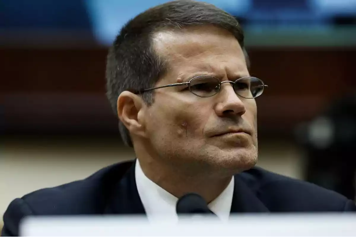 A man with glasses and a dark suit is sitting in front of a microphone in a formal setting. A man with glasses and a dark suit is sitting in front of a microphone in a formal setting.