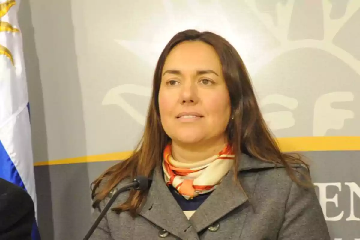 Una mujer con cabello castaño y una bufanda colorida está de pie frente a un micrófono, con una bandera y un fondo gris detrás de ella. Una mujer con cabello castaño y una bufanda colorida está de pie frente a un micrófono, con una bandera y un fondo gris detrás de ella.