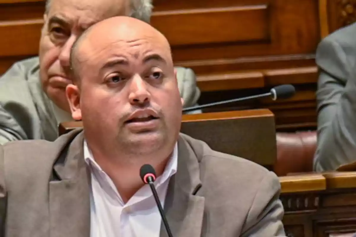 Un hombre hablando en un micrófono en un entorno formal con personas sentadas detrás de él. Un hombre hablando en un micrófono en un entorno formal con personas sentadas detrás de él.