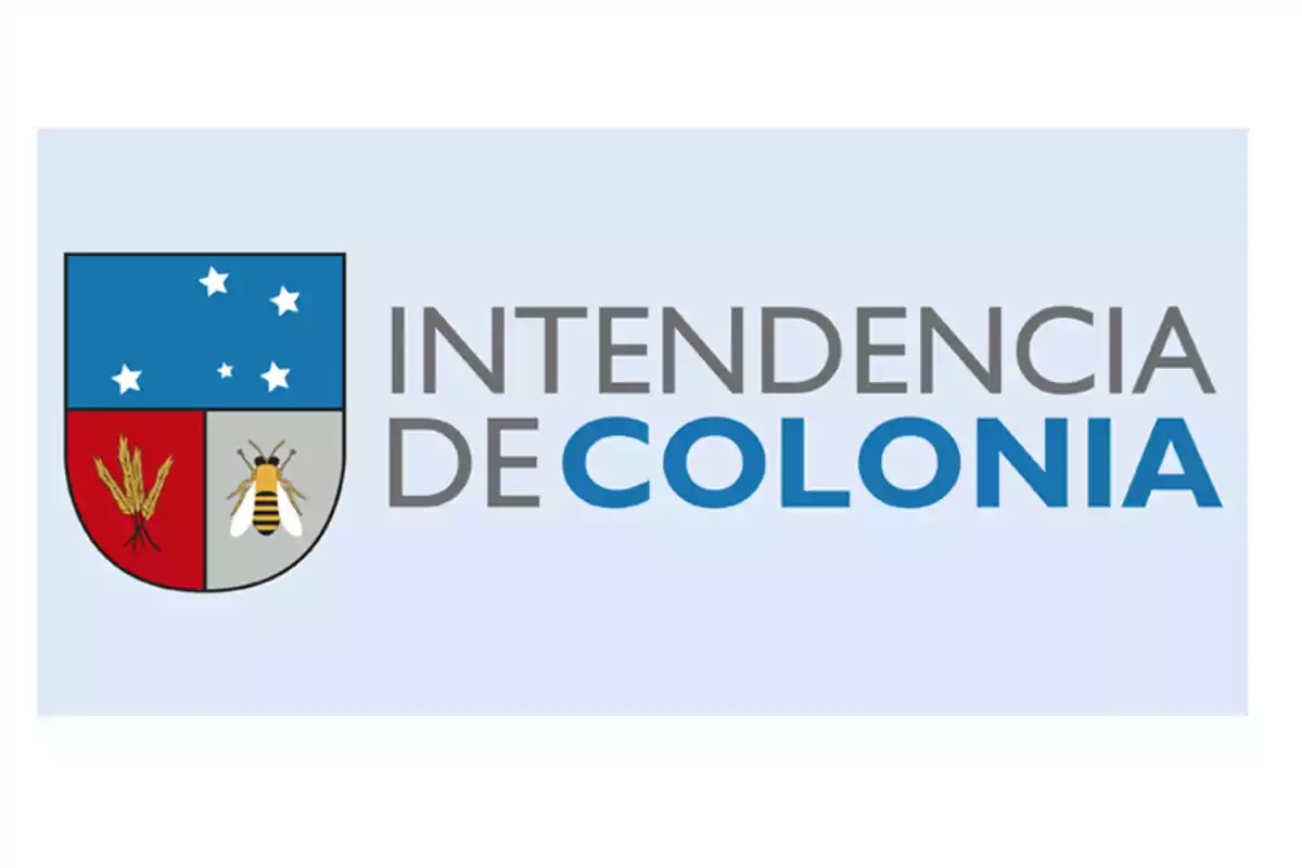 Escudo de la Intendencia de Colonia con espigas de trigo una abeja y estrellas sobre fondo azul junto al texto Intendencia de Colonia. Escudo de la Intendencia de Colonia con espigas de trigo una abeja y estrellas sobre fondo azul junto al texto Intendencia de Colonia.