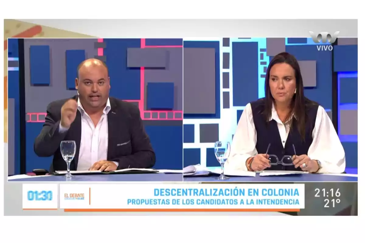 Dos personas participando en un debate televisivo sobre descentralización en Colonia, con gráficos de fondo y texto informativo en la parte inferior.