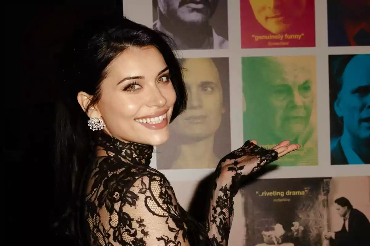 Una persona con un vestido de encaje negro sonríe y señala hacia un collage de imágenes en la pared.