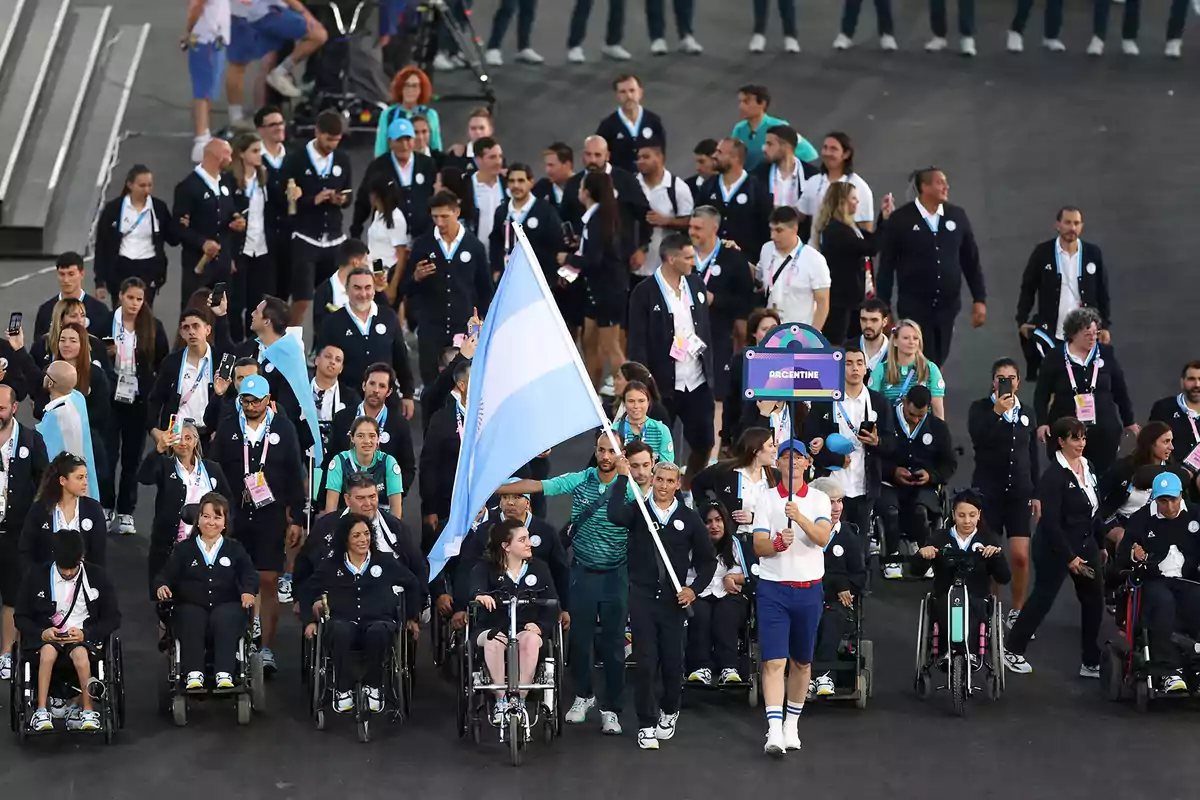 Un grupo de atletas con uniformes oscuros y algunos en sillas de ruedas marchan juntos, llevando una bandera y un cartel que dice 