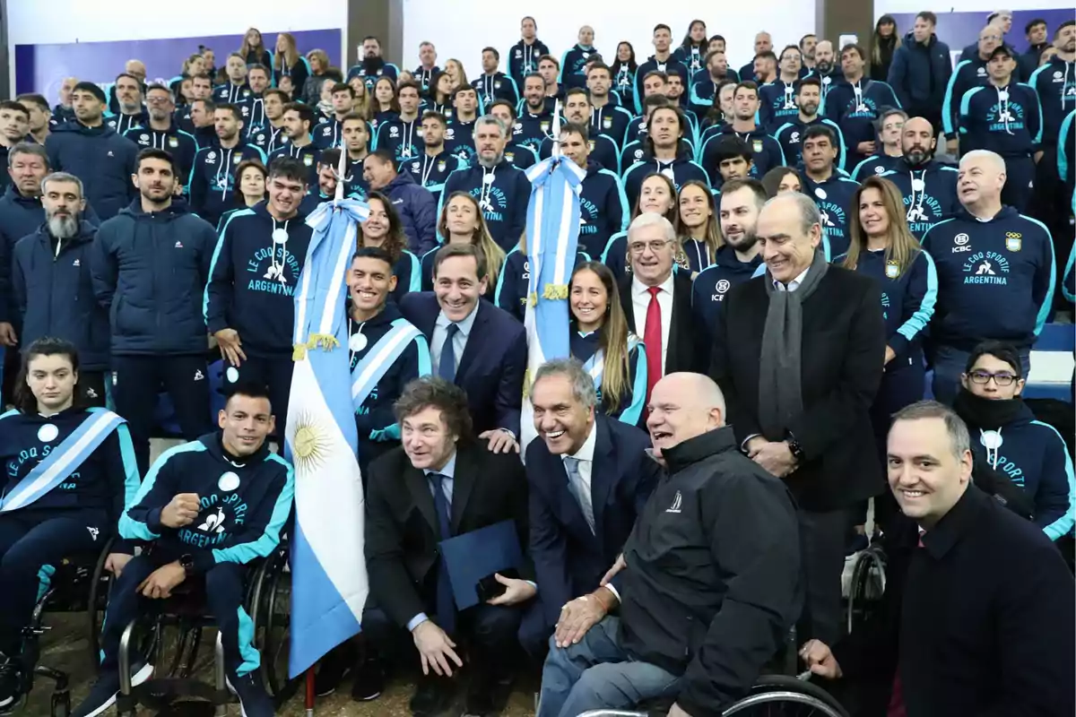 Un grupo de personas, algunas en sillas de ruedas, posando para una foto con banderas argentinas y vestimenta deportiva, acompañados por varias personas en traje.