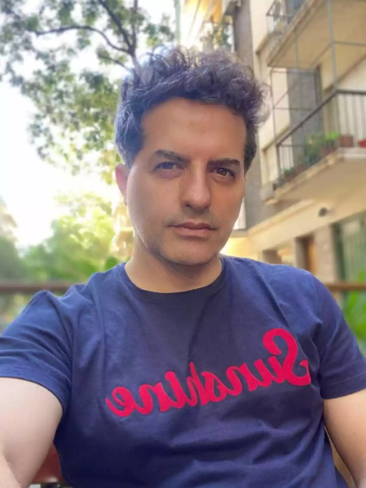 Hombre de cabello rizado y oscuro usando una camiseta azul con la palabra sunshine en letras rojas sentado al aire libre con árboles y edificios de fondo Hombre de cabello rizado y oscuro usando una camiseta azul con la palabra sunshine en letras rojas sentado al aire libre con árboles y edificios de fondo