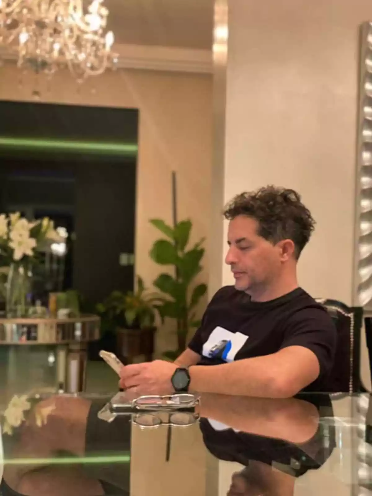 Hombre sentado en una mesa de cristal usando un teléfono móvil en una sala elegante con plantas y un candelabro al fondo Hombre sentado en una mesa de cristal usando un teléfono móvil en una sala elegante con plantas y un candelabro al fondo