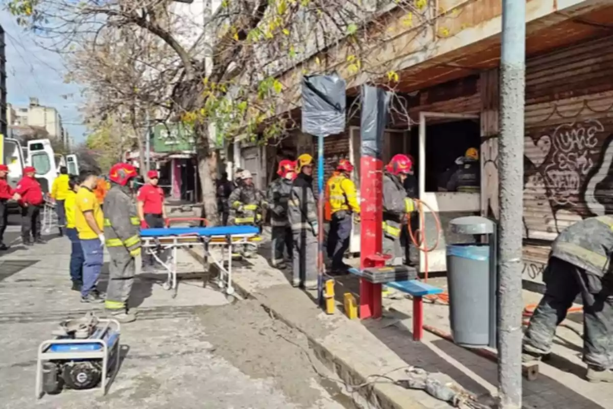 Several firefighters and emergency personnel are working at the entrance of a building with burned metal shutters while a stretcher and rescue equipment are ready on the sidewalk. Several firefighters and emergency personnel are working at the entrance of a building with burned metal shutters while a stretcher and rescue equipment are ready on the sidewalk.