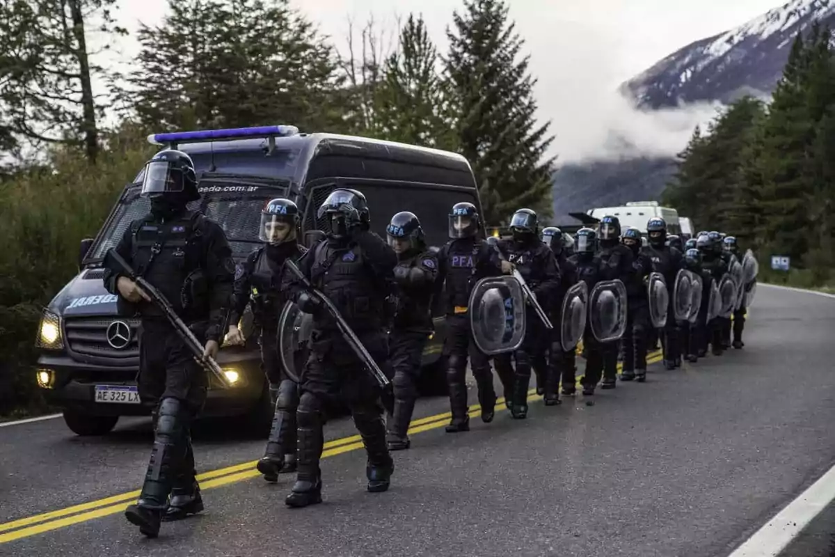 Un grupo de policías antidisturbios camina en formación por una carretera rodeada de árboles y montañas, con un vehículo policial detrás de ellos.