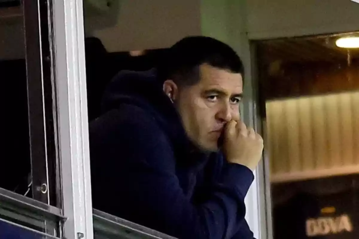 A man with a pensive expression looks out a window while resting his head on his hand.
