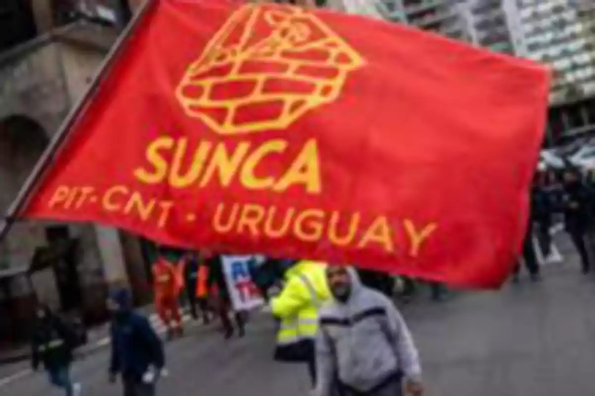 People marching with a red flag that has inscriptions of SUNCA PIT-CNT Uruguay on an urban street.