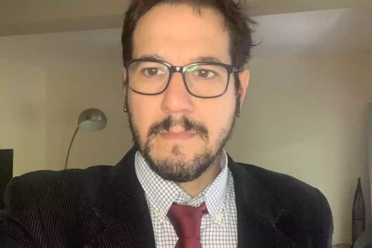 Man with glasses and a beard wearing a dark jacket, checkered jersey, and red tie in a well-lit room