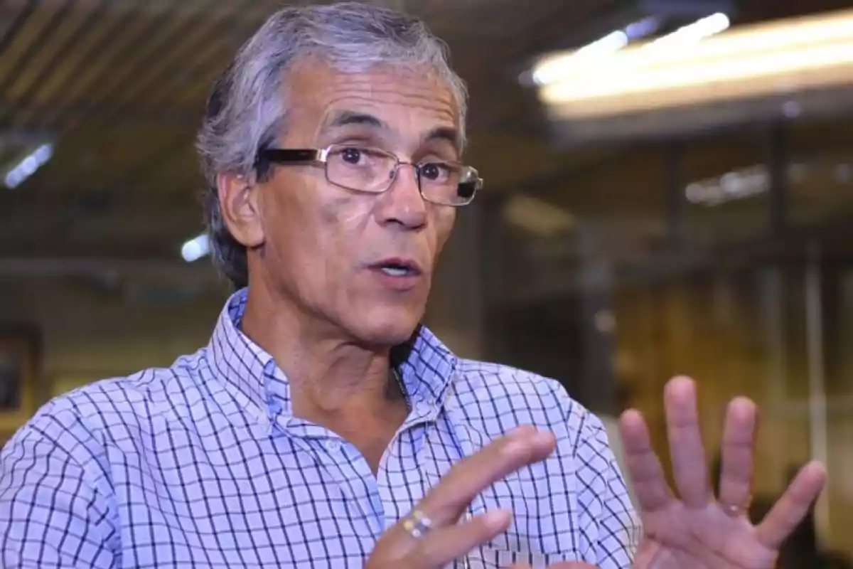 Gray-haired man with glasses speaking and gesturing with his hands, wearing a plaid jersey and in a well-lit indoor setting. Gray-haired man with glasses speaking and gesturing with his hands, wearing a plaid jersey and in a well-lit indoor setting.