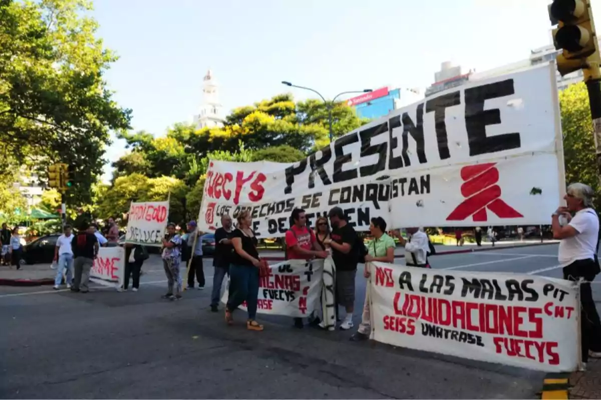 Personas manifestándose en la calle con pancartas grandes que tienen mensajes en letras rojas y negras, rodeados de árboles y edificios al fondo en un día soleado. Personas manifestándose en la calle con pancartas grandes que tienen mensajes en letras rojas y negras, rodeados de árboles y edificios al fondo en un día soleado.