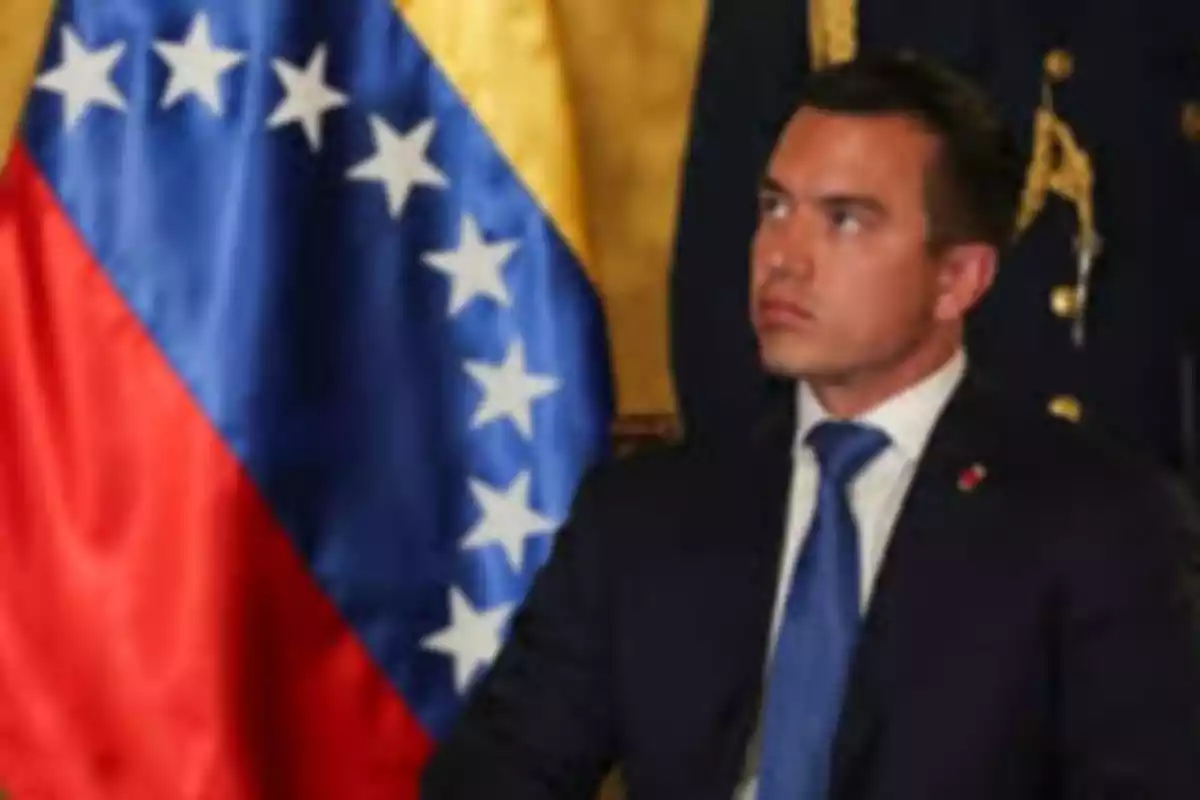 A man in a dark suit and blue tie is sitting in front of a Venezuelan flag.