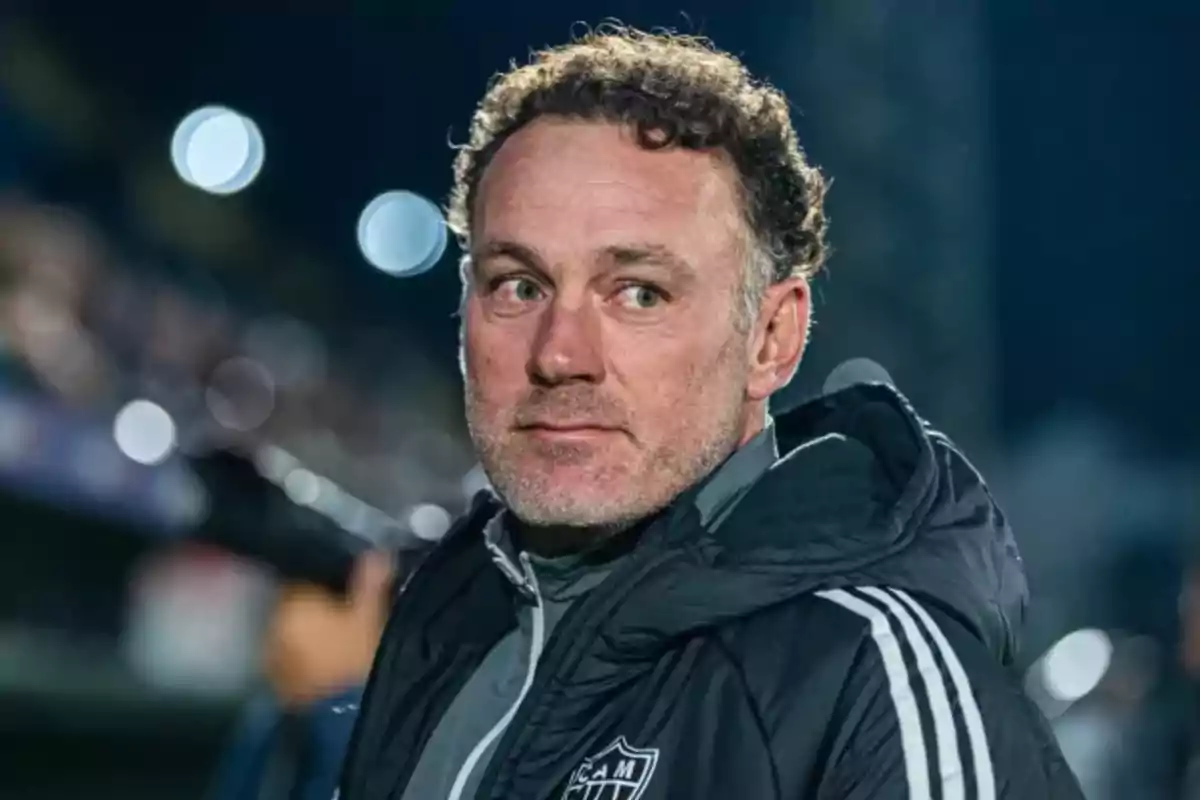 A man in a dark jacket with stubble looks to the side in a nighttime setting with blurred lights in the background.