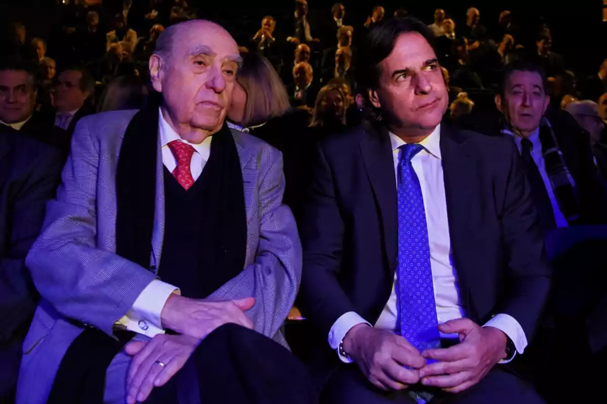 Dos hombres sentados en un evento formal, uno con traje gris y corbata roja, el otro con traje oscuro y corbata azul, rodeados de otras personas en un auditorio. Dos hombres sentados en un evento formal, uno con traje gris y corbata roja, el otro con traje oscuro y corbata azul, rodeados de otras personas en un auditorio.