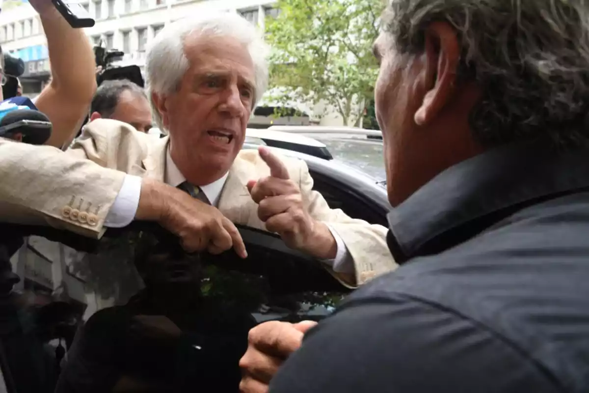 Un hombre de cabello canoso y traje claro gesticula con intensidad mientras habla con otra persona en la calle rodeado de cámaras y micrófonos. Un hombre de cabello canoso y traje claro gesticula con intensidad mientras habla con otra persona en la calle rodeado de cámaras y micrófonos.