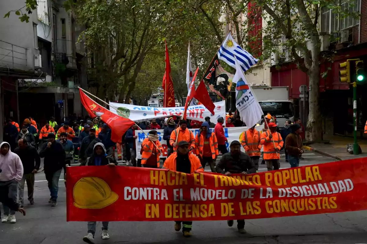 Workers' demonstration with helmets and orange vests marching down a street, carrying a red banner with yellow and white letters, several flags, and trees on the sides