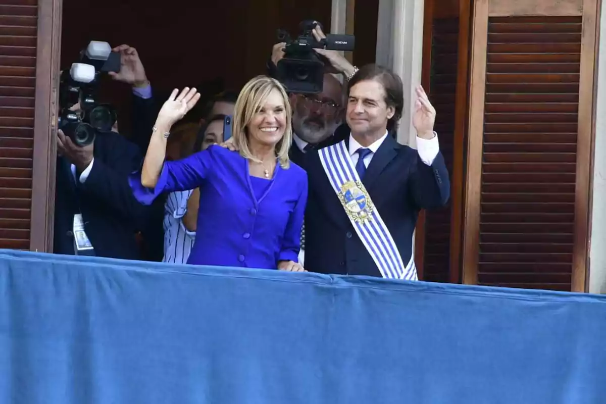A woman and a man dressed formally wave and smile from a balcony while several people take photos and record them.
