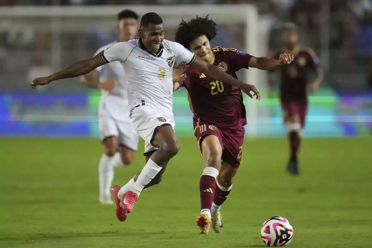 Two players compete for the ball during a match, one wearing a white uniform and the other wearing a burgundy uniform, both showing physical effort in the play. Two players compete for the ball during a match, one wearing a white uniform and the other wearing a burgundy uniform, both showing physical effort in the play.