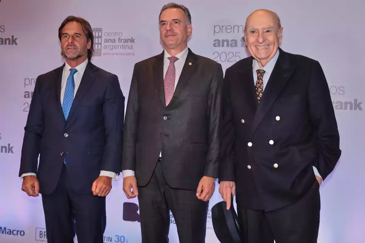 Three men dressed in suits pose smiling in front of a backdrop with the Centro Ana Frank Argentina logo during a formal event.